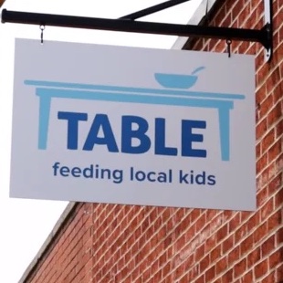 Hanging sign with logo and text: TABLE feeding local kids